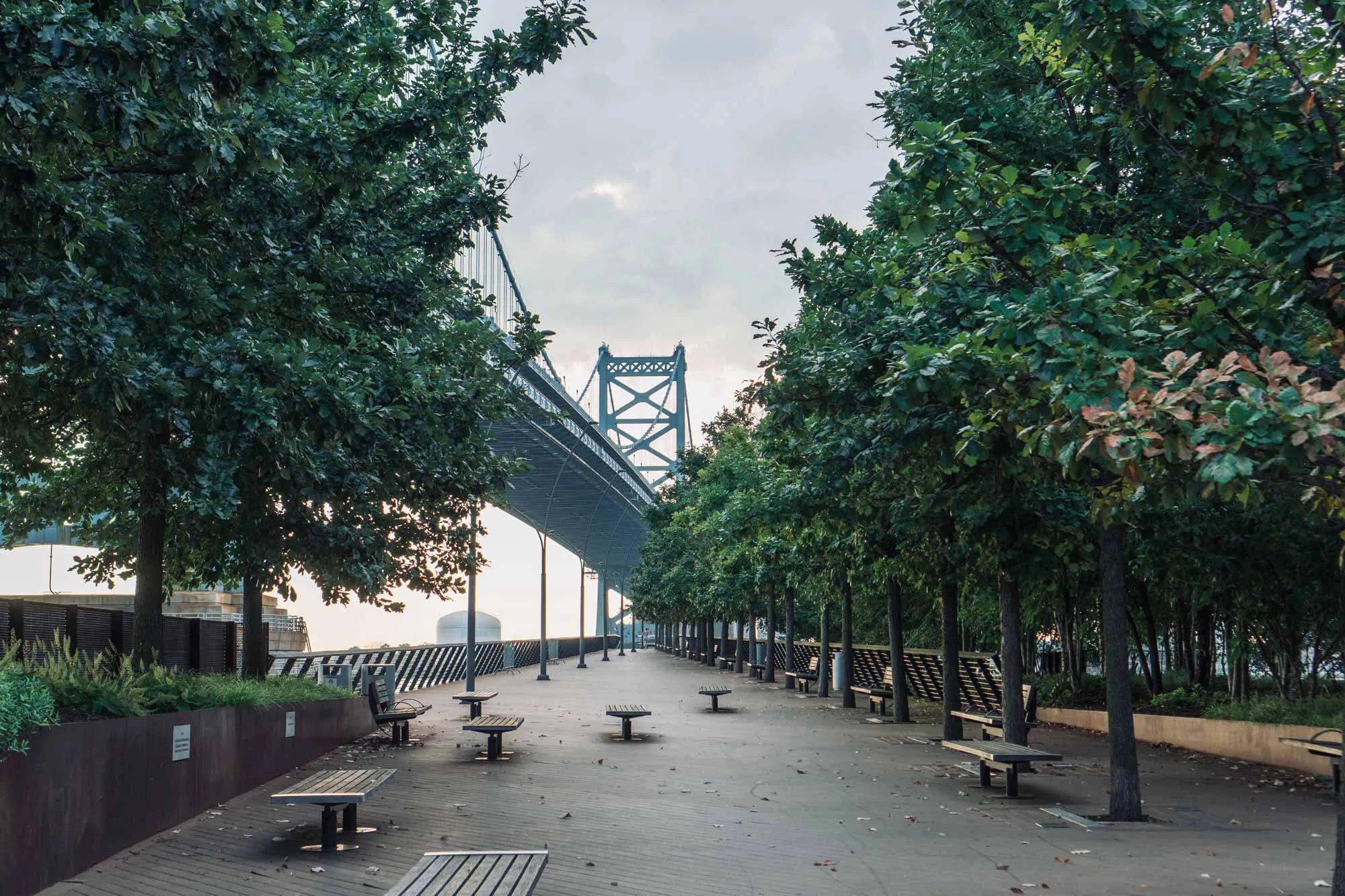 Benches line a sidewalk in a seaside park, offering a view of a bridge in the distance.